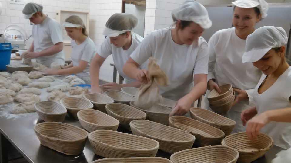 Vpředu se moučí ošatky, děvčata zpracovávají těsto na žitno-pšeničný chléb Šumava